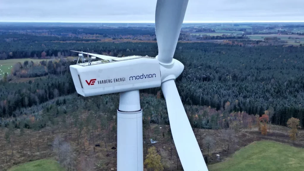 Modvion Modulare Windkrafttürme aus Holz 1 Luftaufnahme einer Modvion-Holzwindturbine mit Logos von Varberg Energi und Modvion auf der weißen Gondel, im Hintergrund schwedische Wald- und Wiesenlandschaft