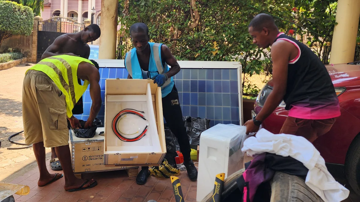 Nigerias Solarboom in Onitsha: Drei Männer packen vor einem Wohnhaus Lithiumbatterie und Kabel für eine Solaranlage aus, dahinter Solarmodule
